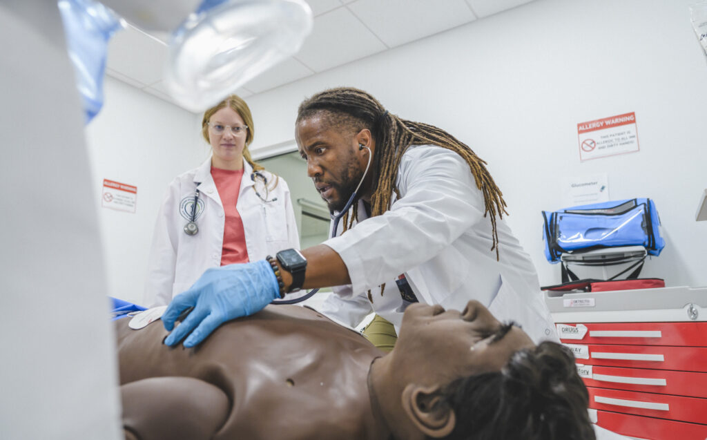 WAUSM medical students in white coats practice a procedure on the Apollo patient simulator in a WAUSM simulation lab, with a vitals monitor behind them.