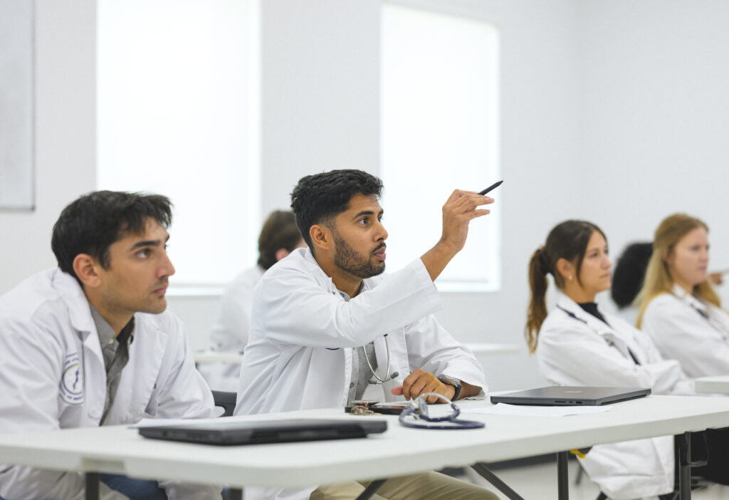 WAUSM medical students in white coats sit at classroom tables during an IPE patient-clinic debrief session, one student speaking while others listen.